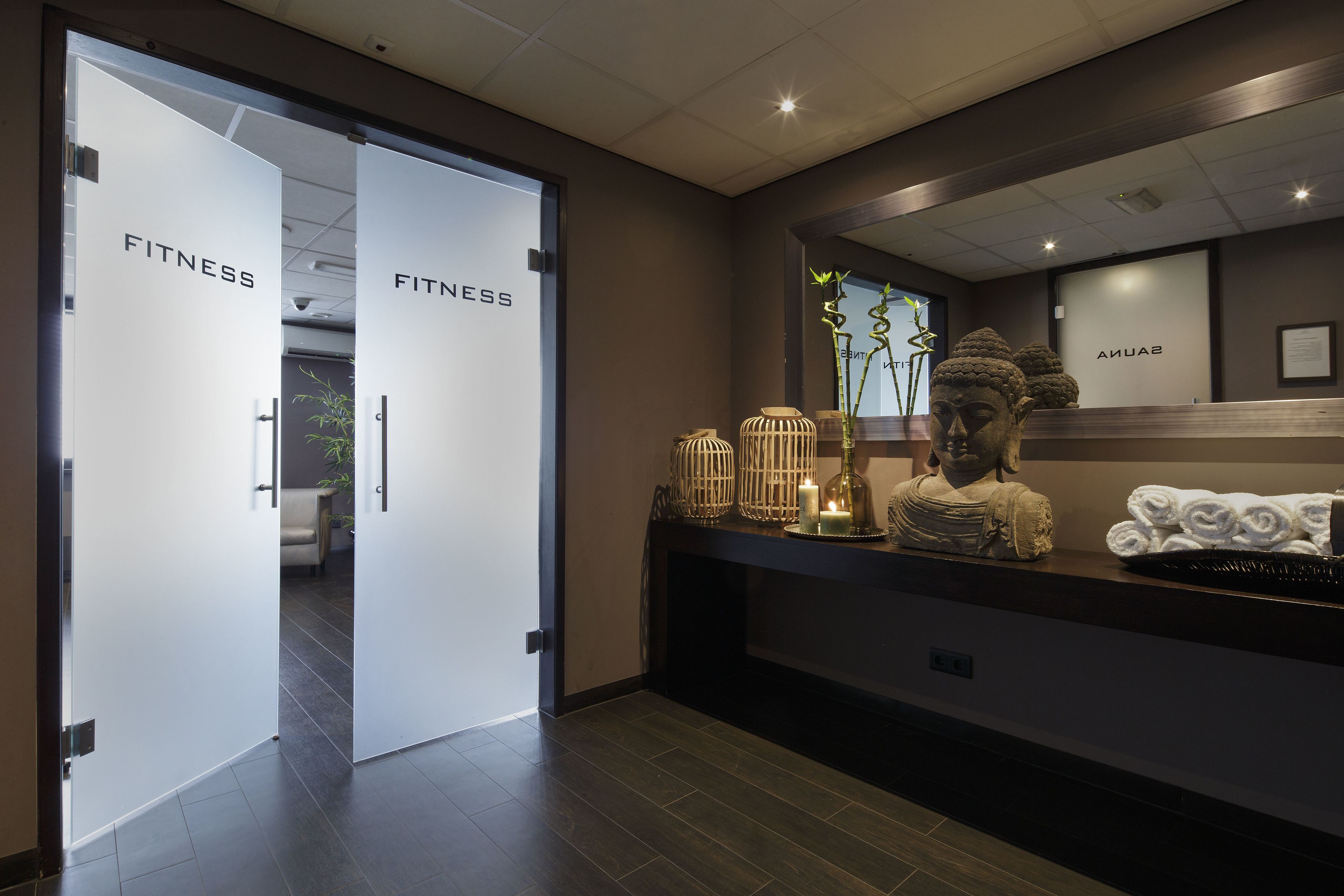 Et receptionsområde med to døre mærket 'FITNESS', en receptionsdisk med dekorative genstande og en statue.