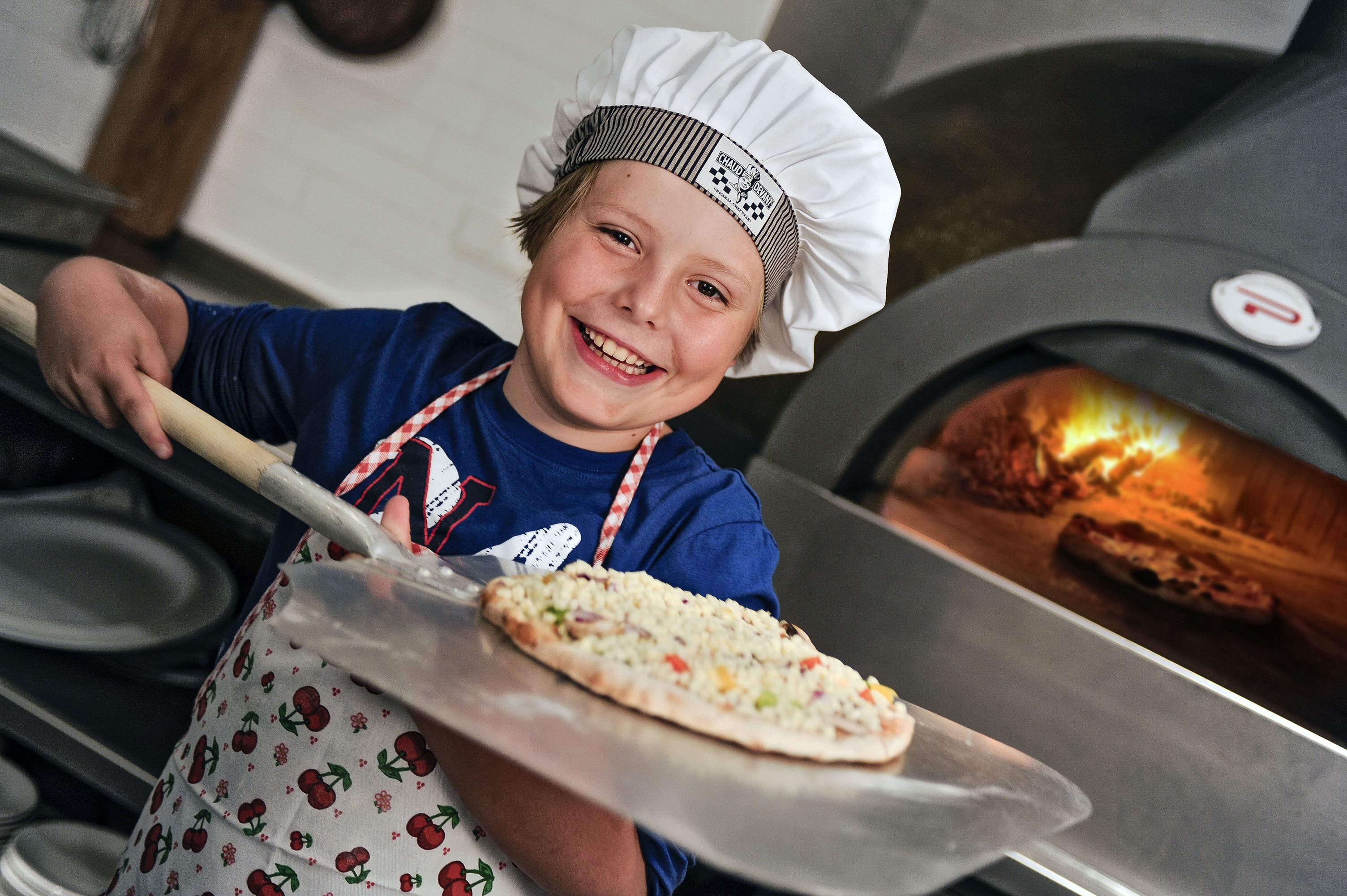 A person in a chef's hat holding a pizza with a pizza oven in the background.