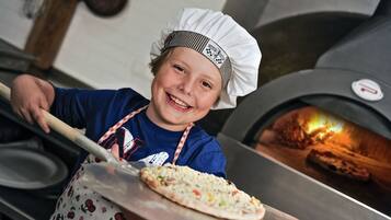 A person in a chef's hat holding a pizza with a pizza oven in the background.