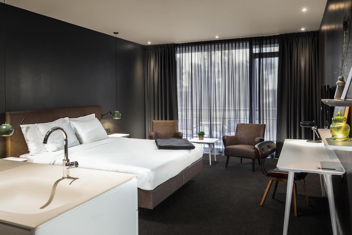 Una habitación de hotel moderna con una cama grande, un escritorio, dos sillas, una lámpara de escritorio, una mesita pequeña y un jarrón decorativo.