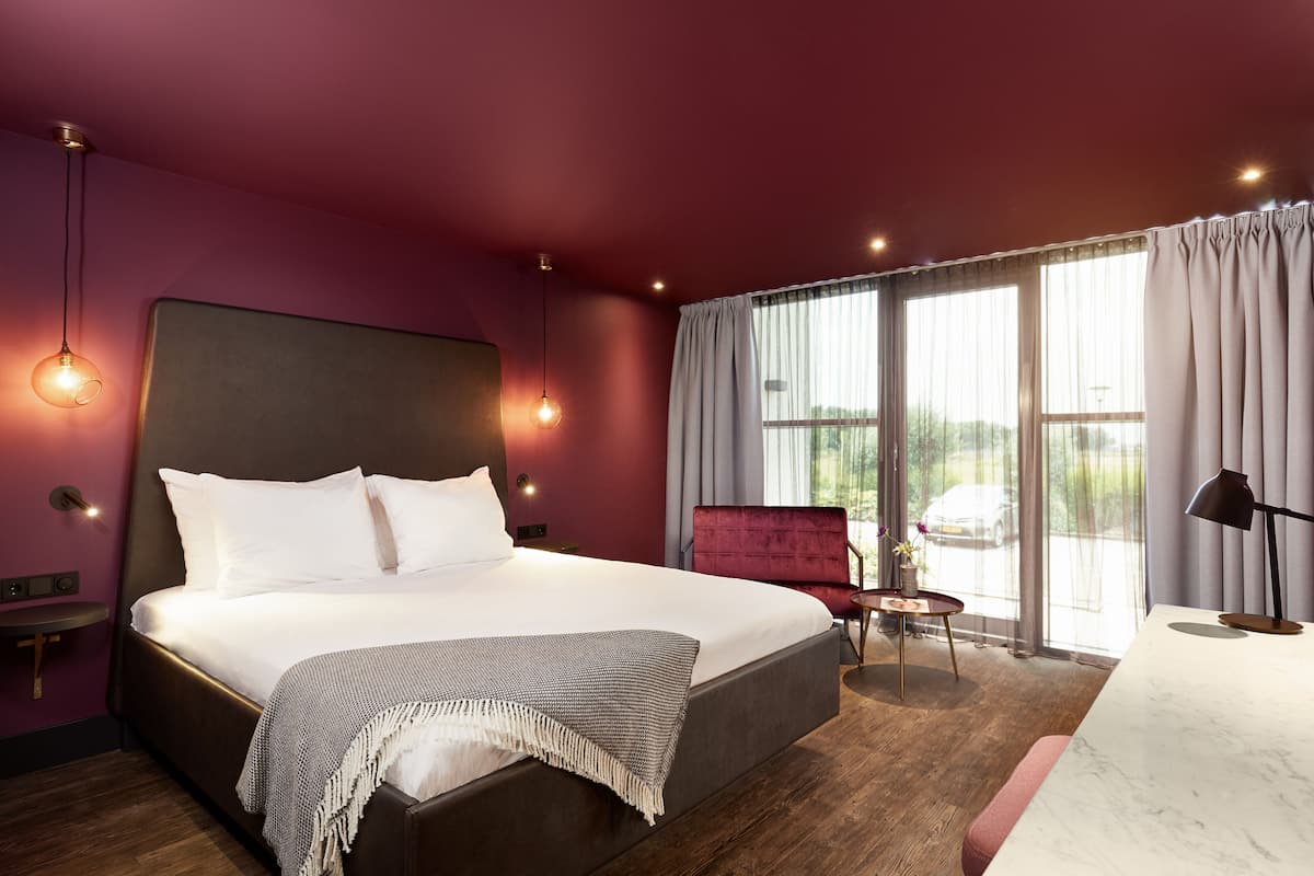 Habitación de hotel moderna con una cama grande, una silla roja, una mesita pequeña y vistas al exterior.