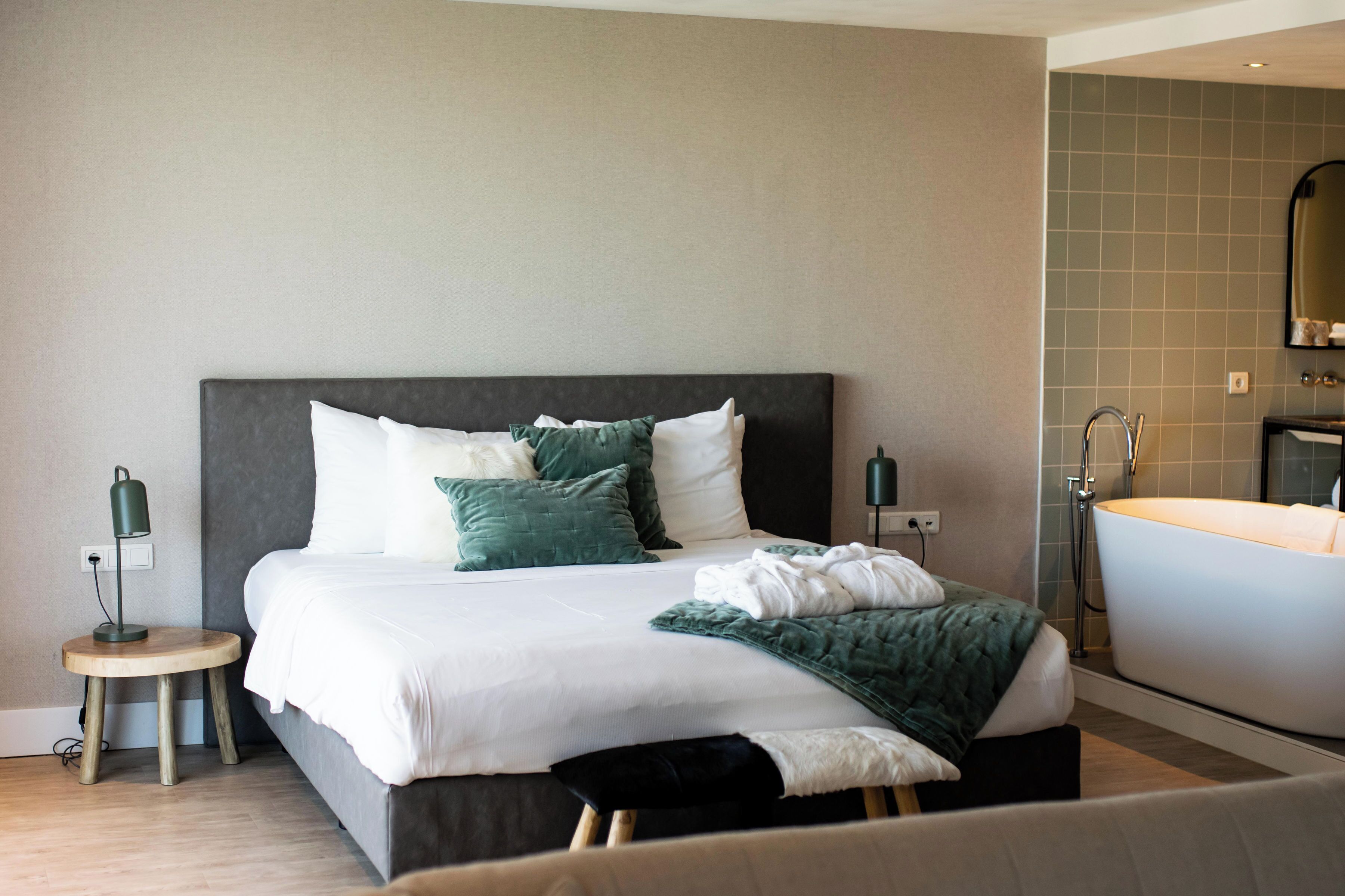 Una habitación de hotel moderna con una cama grande, una mesita de noche con lámpara, un baño con bañera y un espejo en la pared.