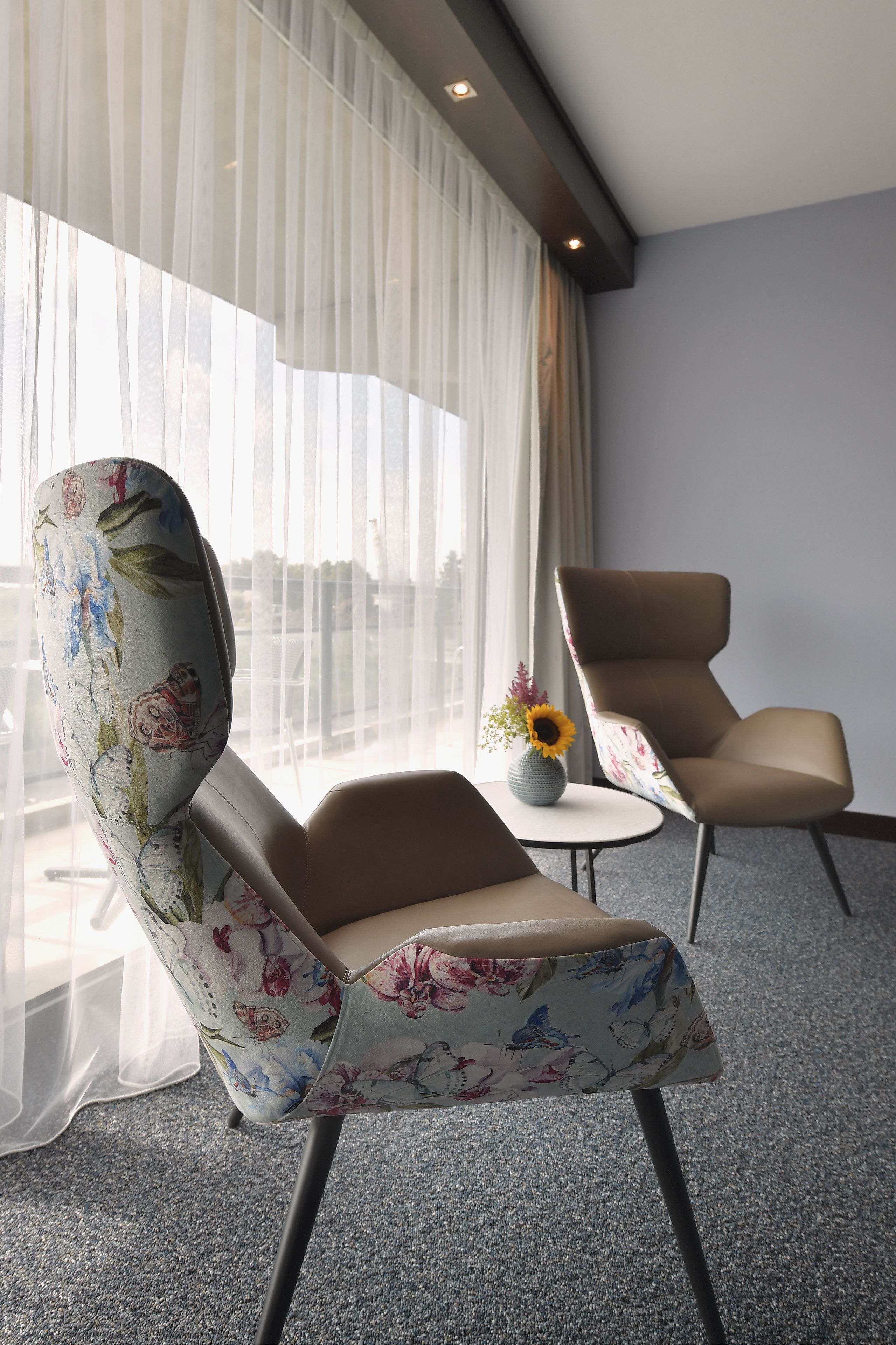 Une chambre d’hôtel moderne avec une grande fenêtre, un fauteuil à motifs floraux, un fauteuil uni et une petite table sur laquelle est posé un vase de fleurs.