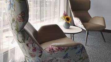 Une chambre d’hôtel moderne avec une grande fenêtre, un fauteuil à motifs floraux, un fauteuil uni et une petite table sur laquelle est posé un vase de fleurs.