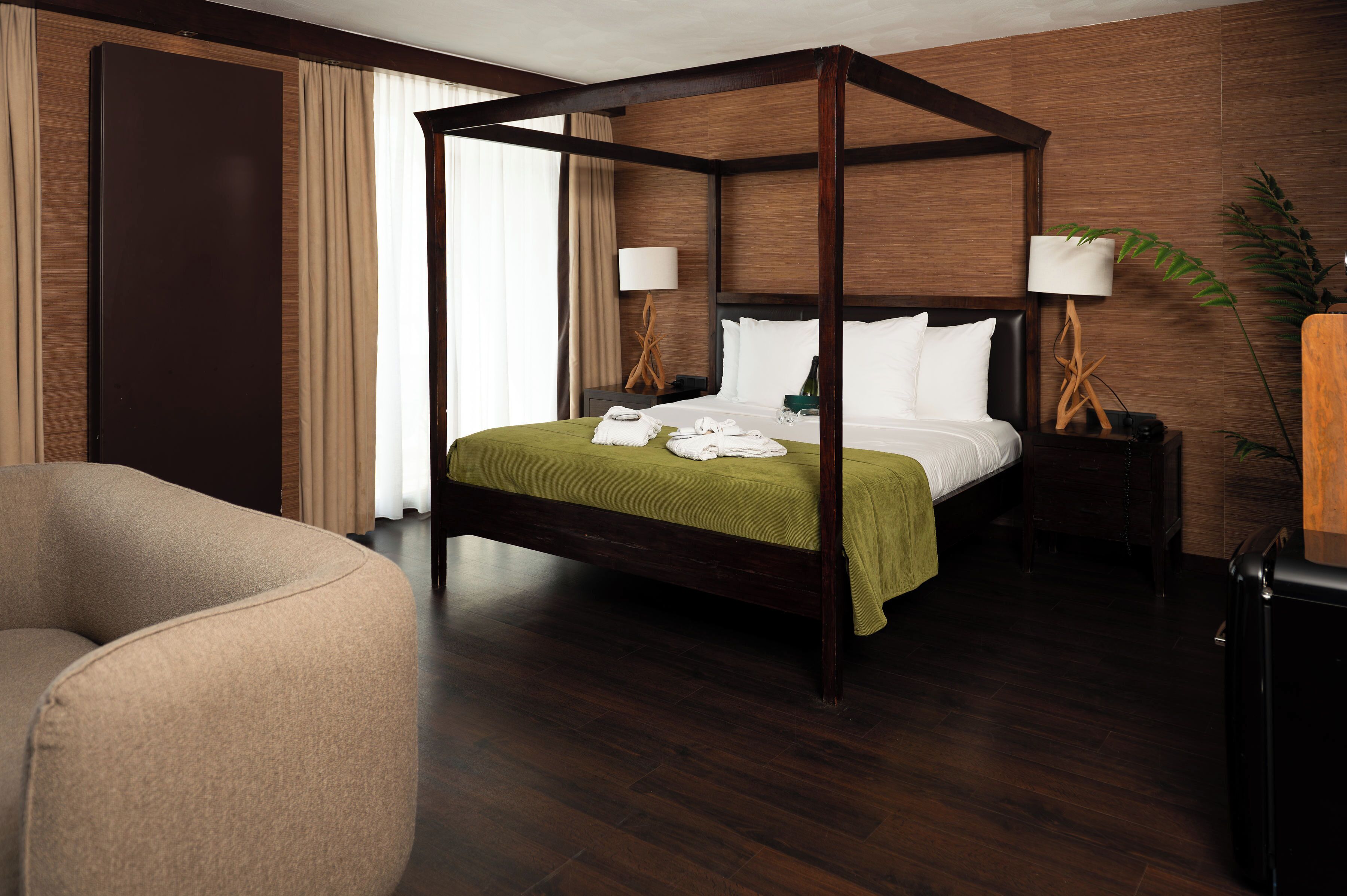 A four-poster bed with a green bedspread, two bedside tables with lamps, a dark wooden floor, and a large window with curtains.