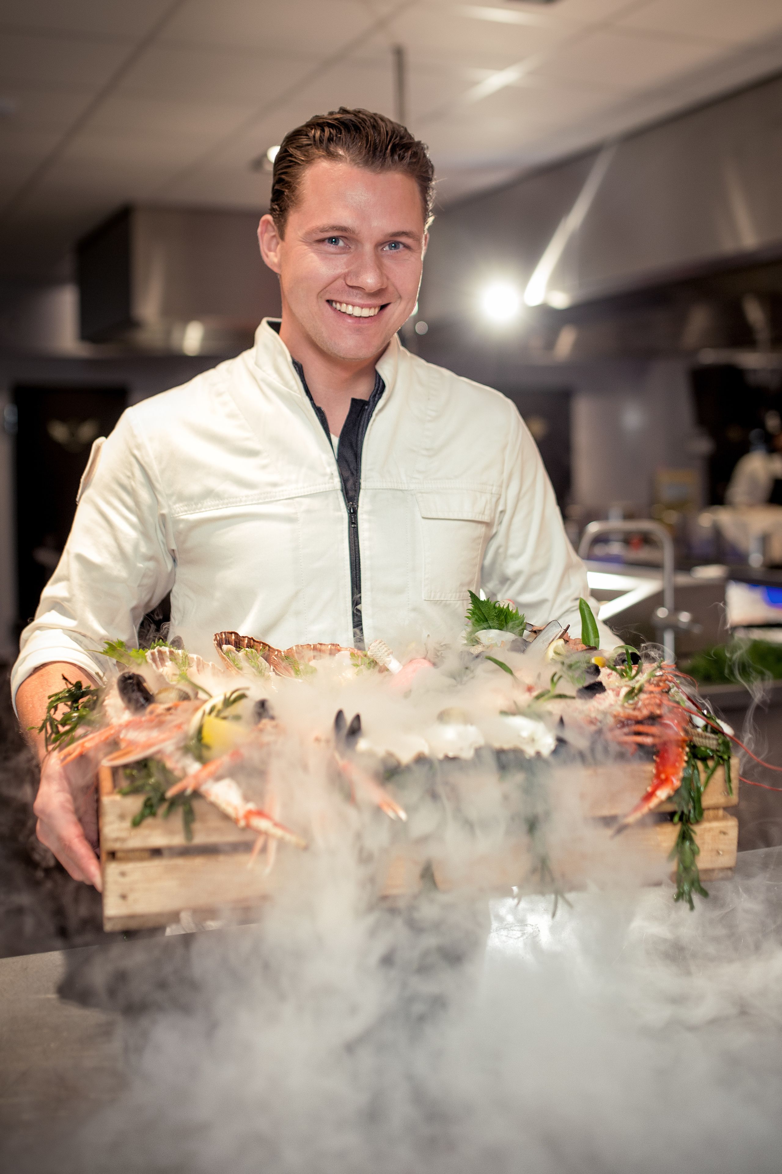 A chef holding a wooden crate filled with seafood, steam rising from the contents.