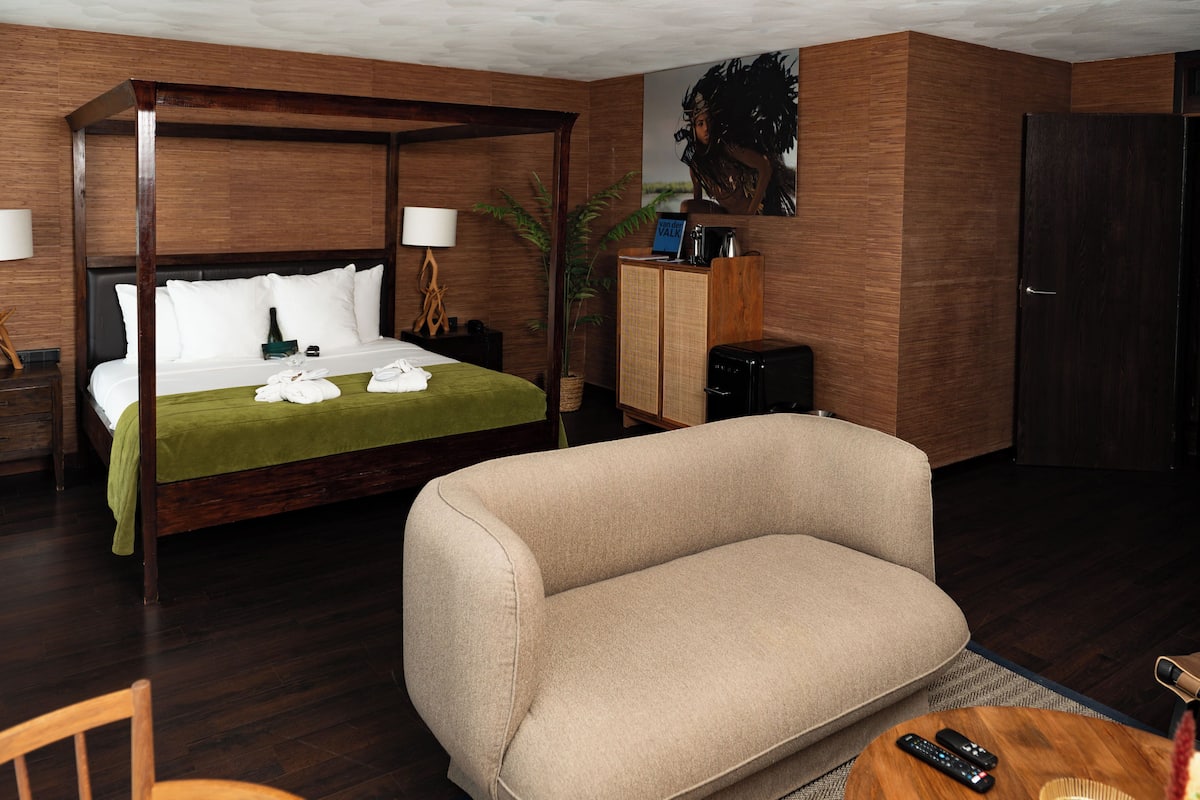 Habitación de hotel con cama con dosel, un sofá, un escritorio con lámpara, cafetera y televisor.