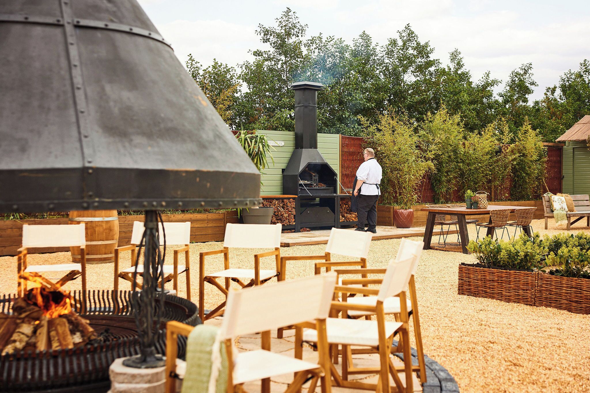 Un patio con chimenea de leña, mobiliario exterior y una valla verde al fondo.