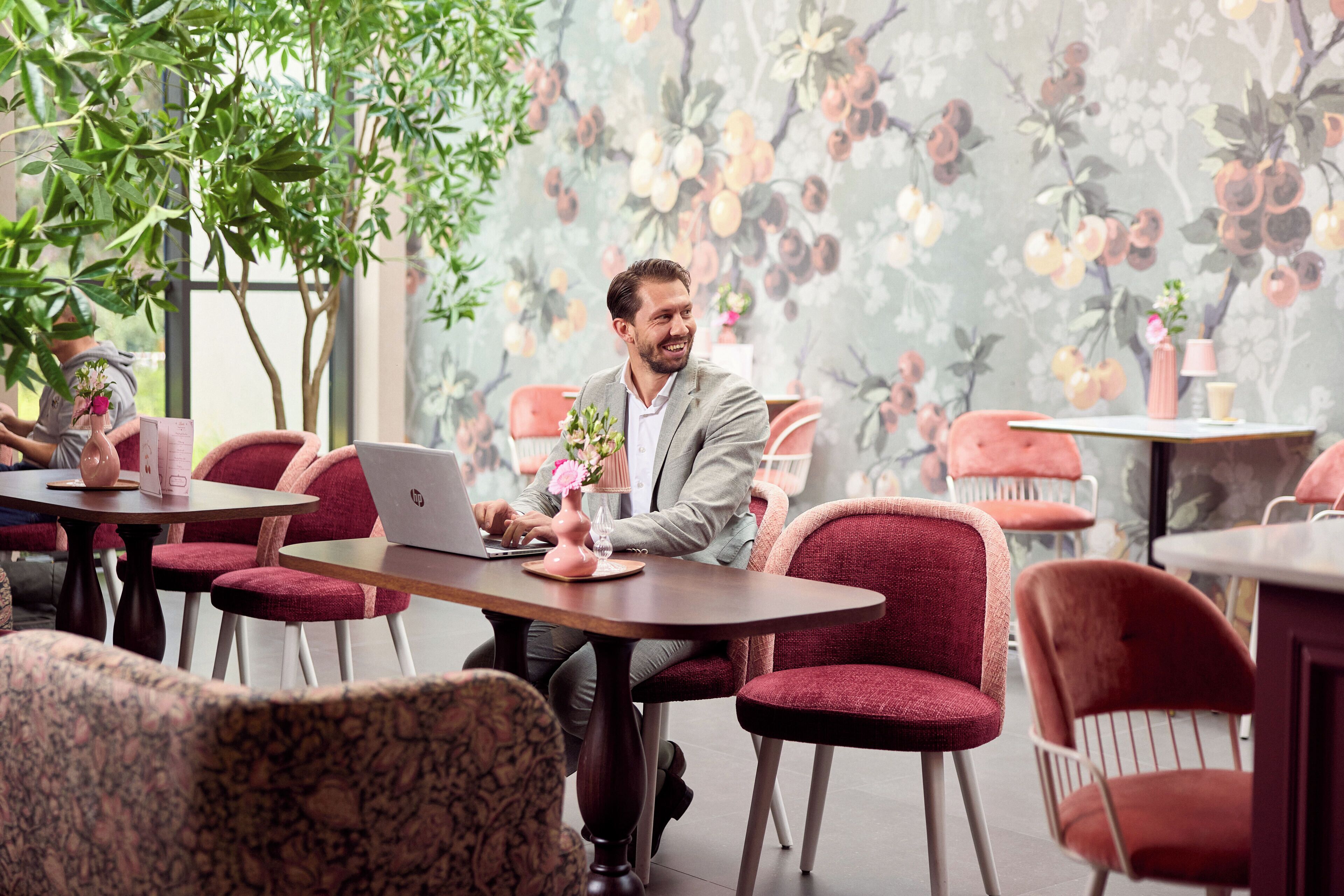 A man sitting at a table with a laptop, surrounded by red chairs and a floral wallpaper.