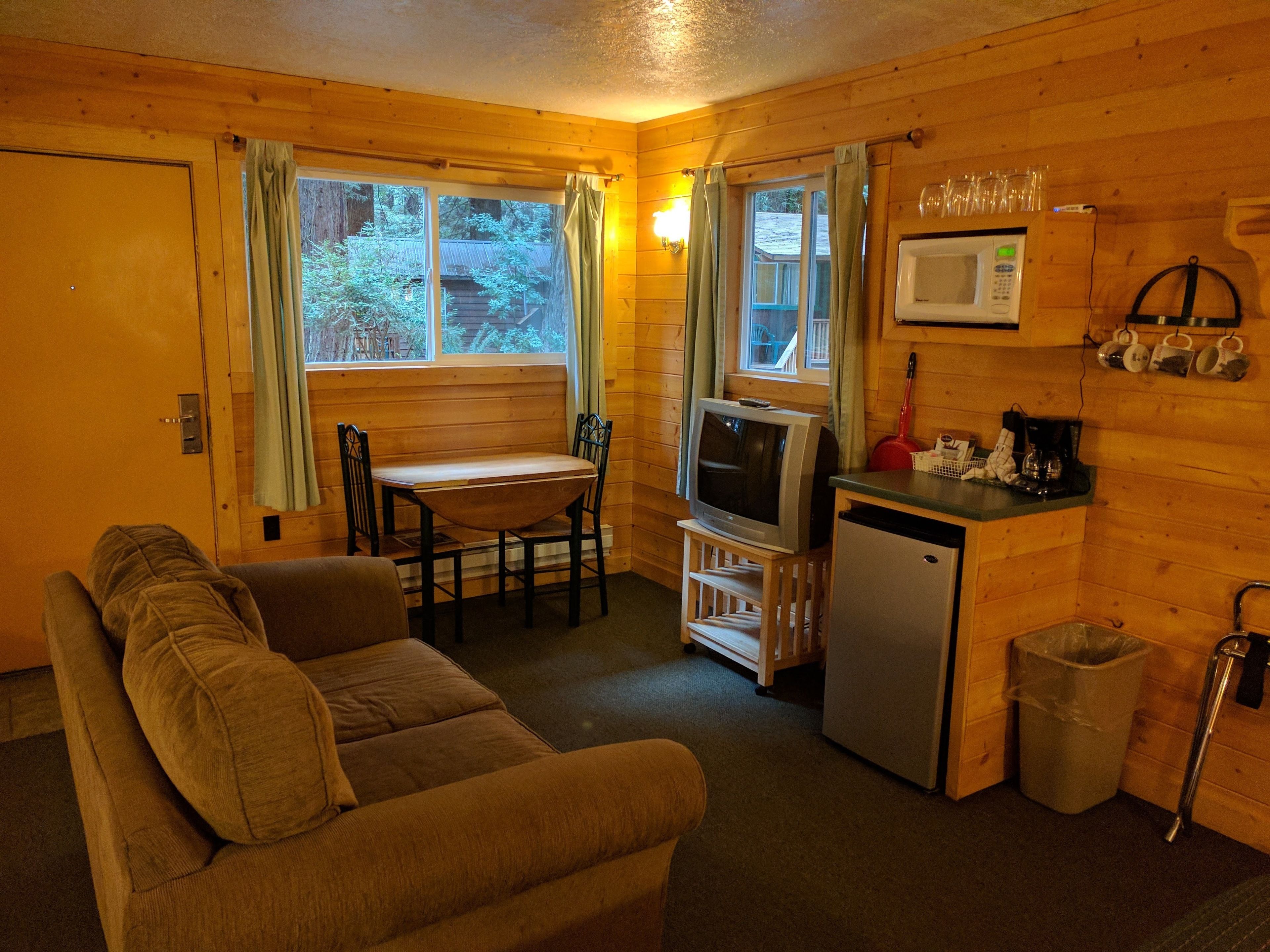 A cozy cabin interior with a wooden interior, a dining table with chairs, a sofa, a kitchenette with a microwave, and a trash bin.