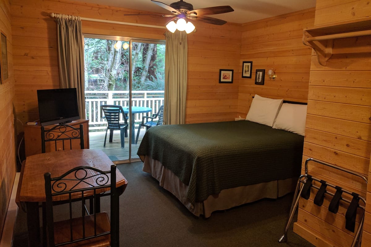 A wooden cabin room with a bed, a desk with a chair, a television, and a balcony with a table and chairs.