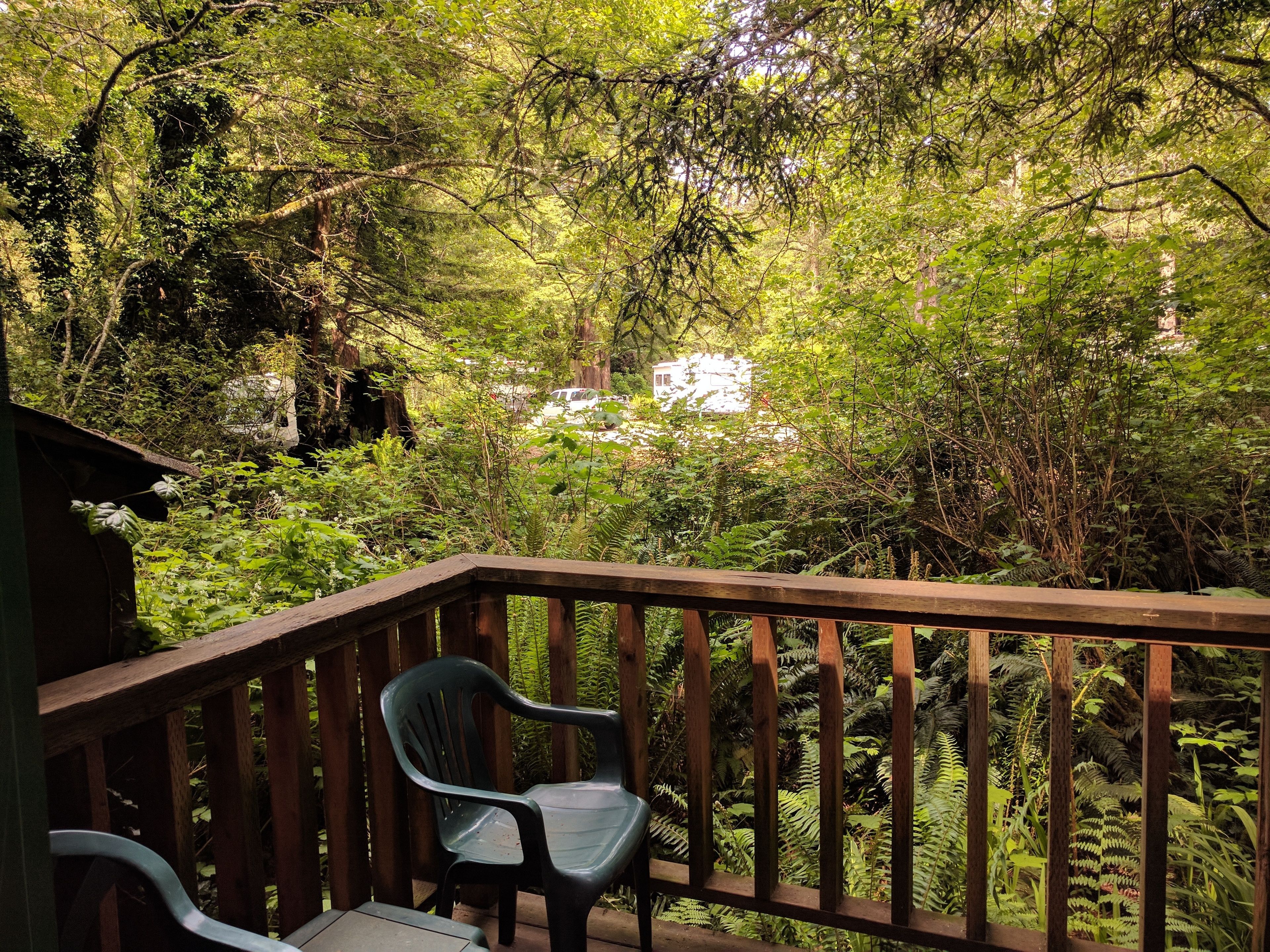 A wooden deck with two plastic chairs, surrounded by dense green foliage and a white RV in the background.