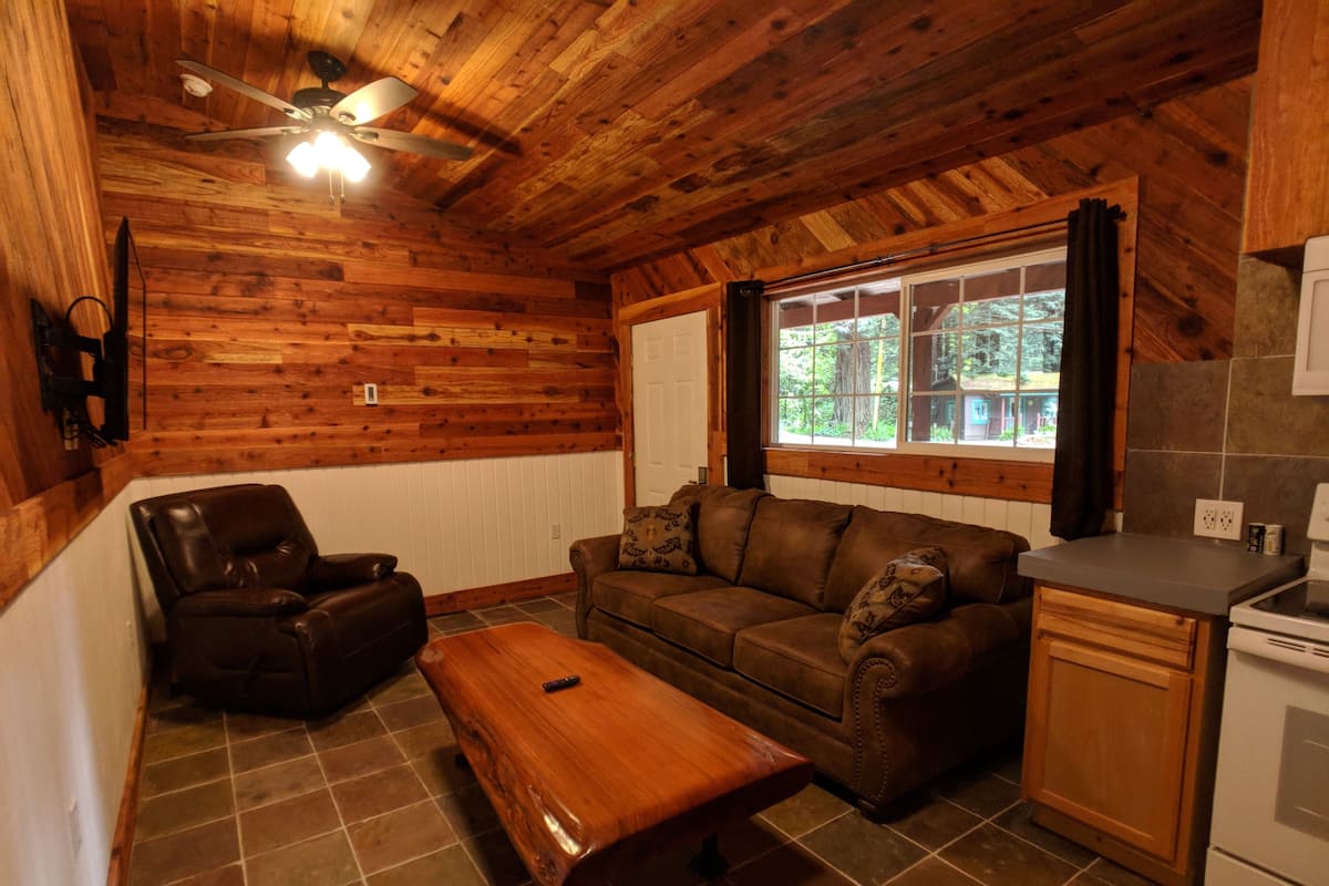 A cozy cabin interior with a wooden ceiling, a leather chair, a sofa, a wooden table, and a kitchen area with a window.