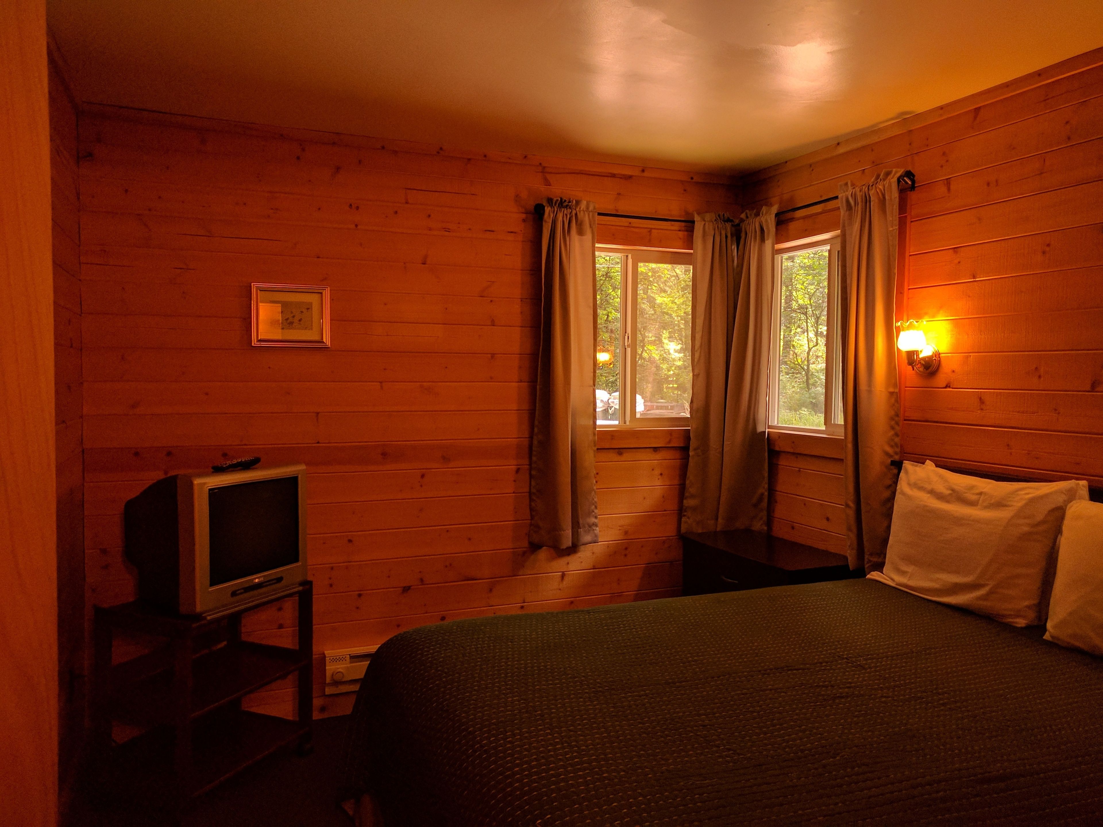 A wooden cabin room with a bed, a television on a stand, and a window with curtains.