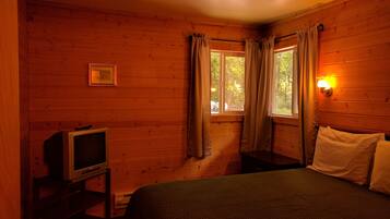 A wooden cabin room with a bed, a television on a stand, and a window with curtains.