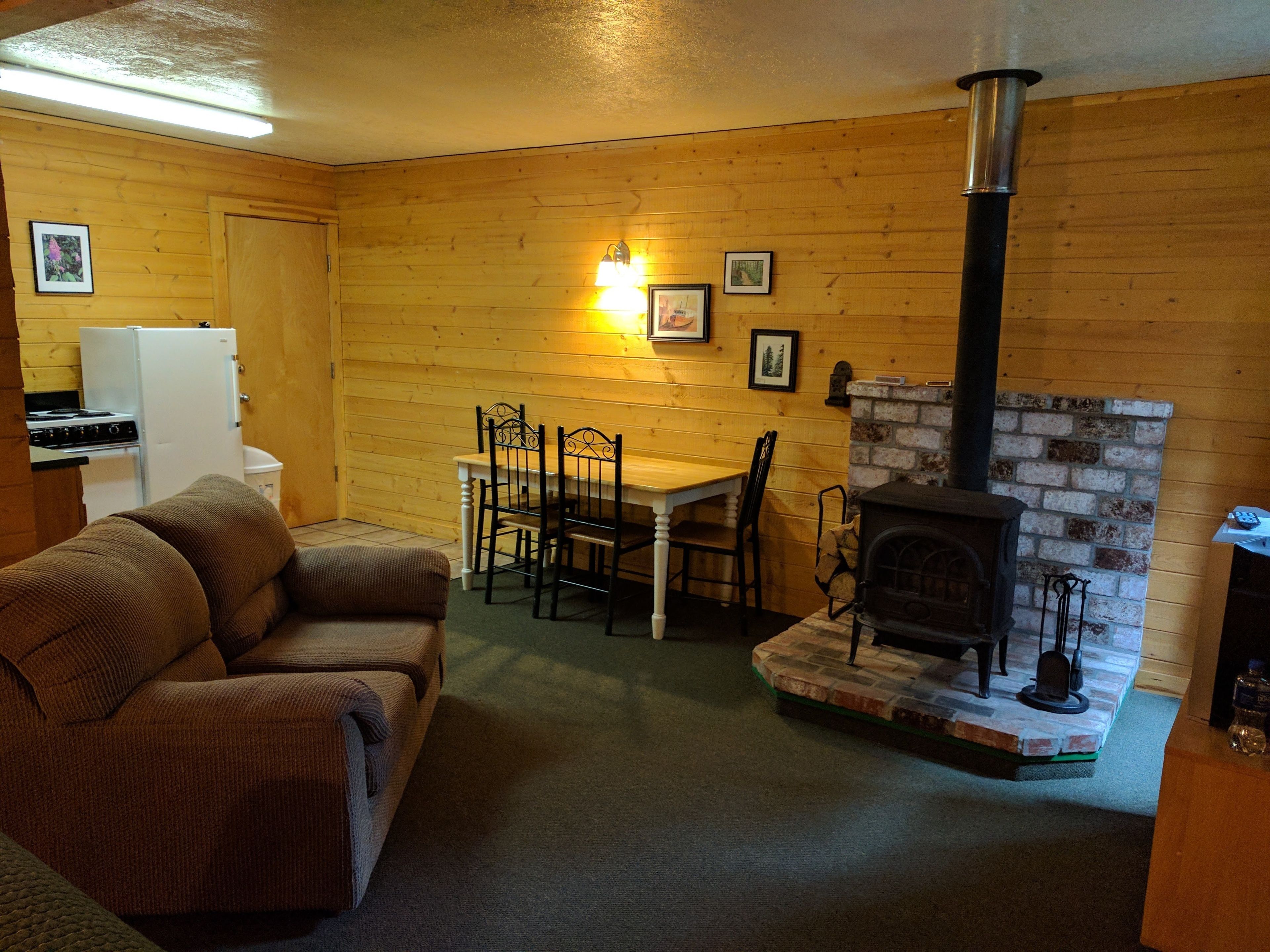 A cozy living room with a fireplace, wooden walls, a dining table with chairs, a sofa, and a refrigerator.