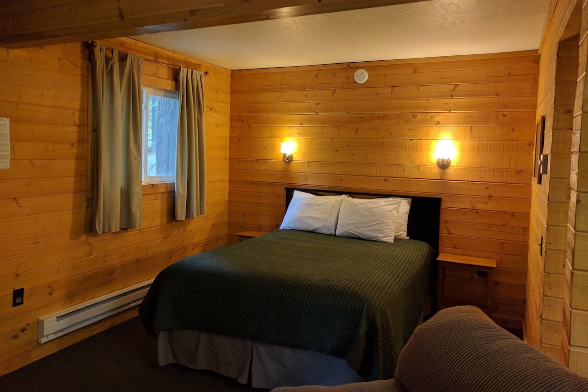 A wooden-paneled room with a bed, two wall-mounted lights, a window with curtains, and a thermostat.
