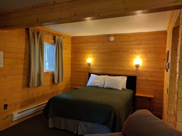 Una habitación con paneles de madera, una cama, dos luces fijadas a la pared, un ventanal con cortinas y un termostato.