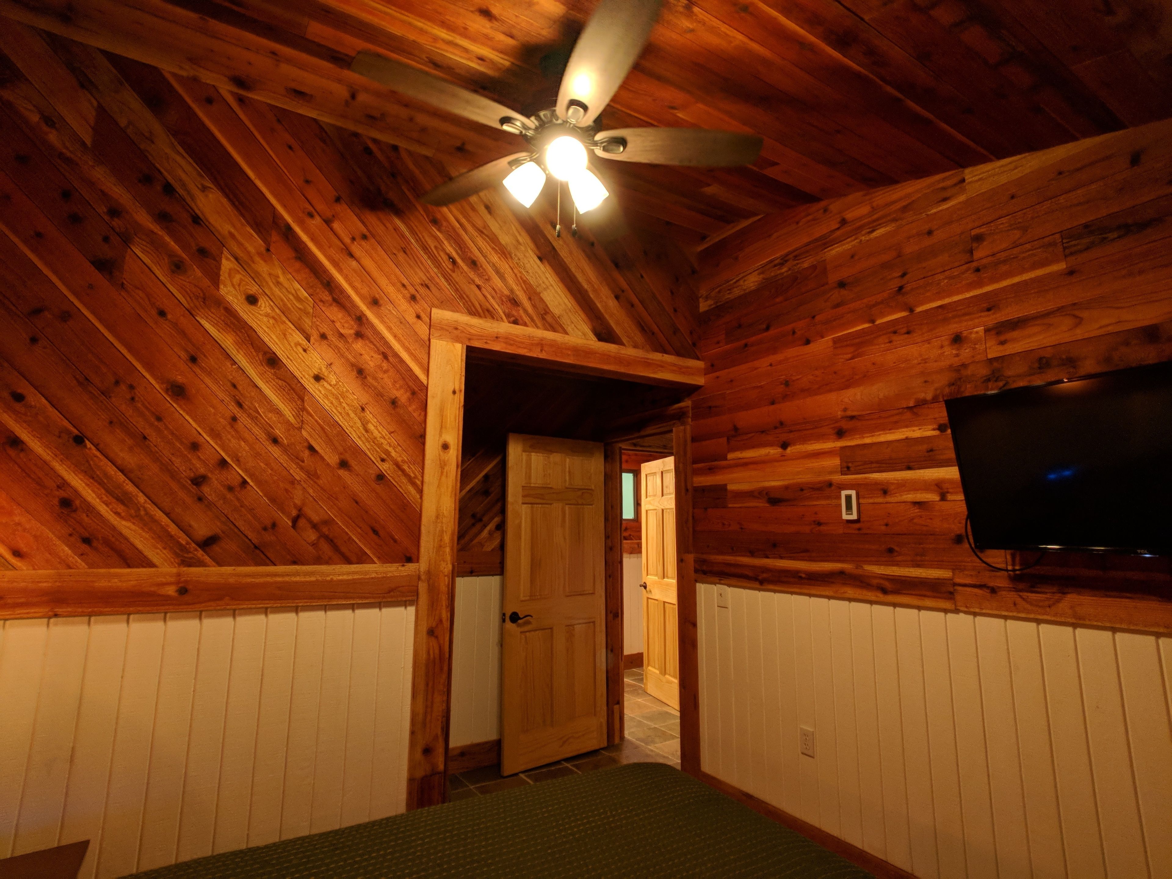 A room with wooden walls and a sloped ceiling, a door leading to another room, and a flat-screen TV mounted on the wall.