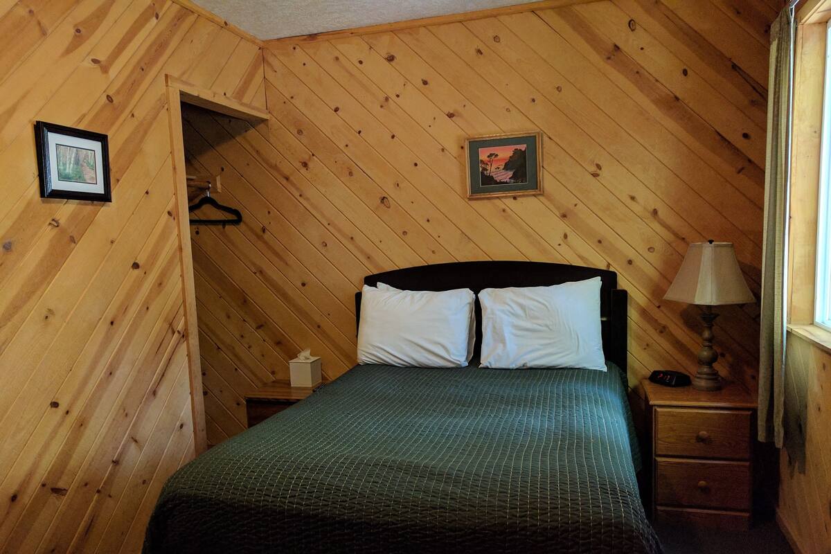 A wooden-paneled bedroom with a bed, two pillows, a nightstand, a lamp, and a framed picture on the wall.