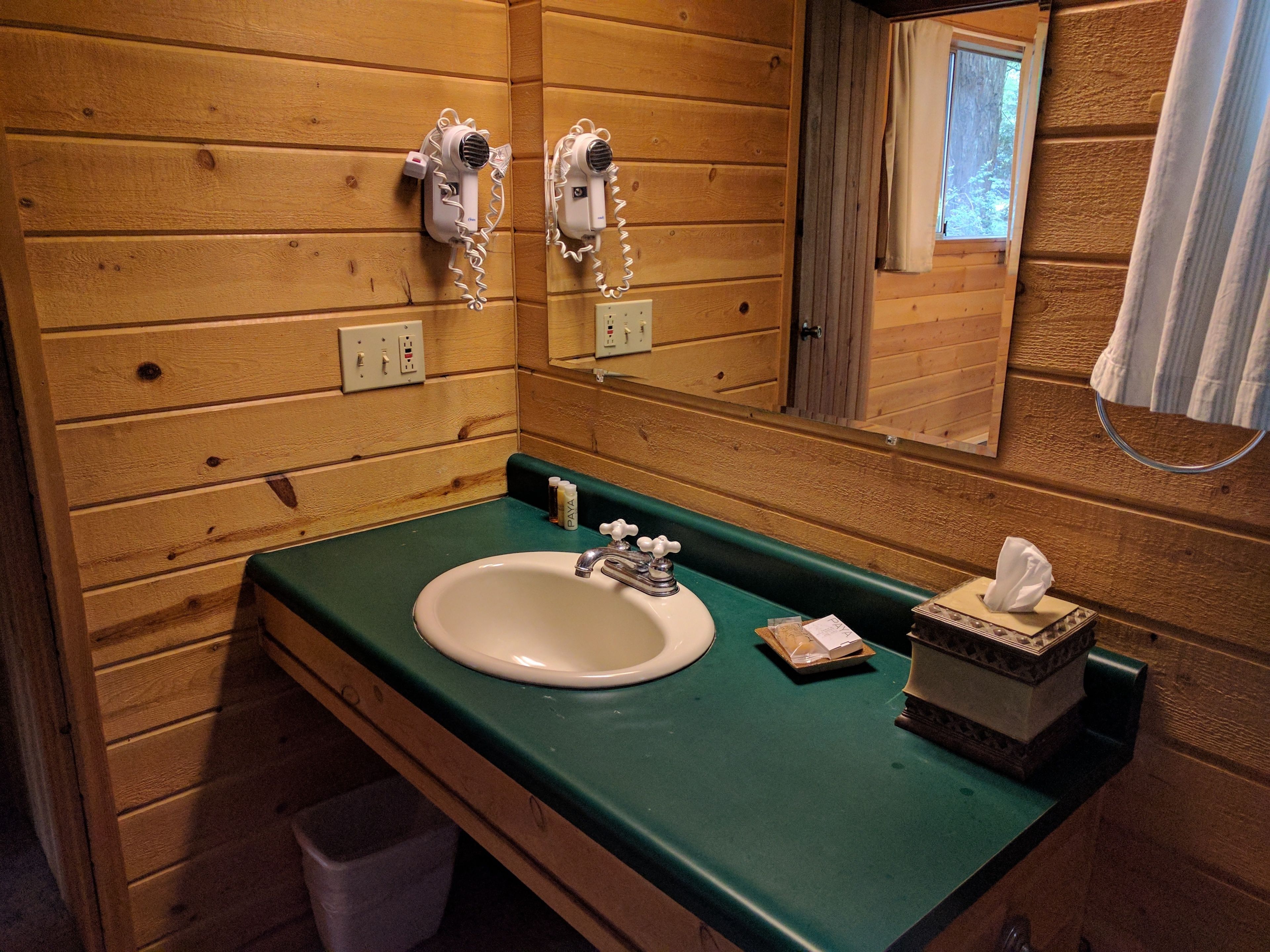 Casa de banho de cabana de madeira com bancada verde, uma pia branca, um espelho e dois secadores de cabelo fixados na parede.