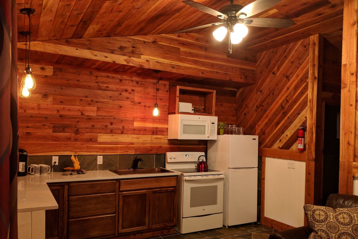 A compact kitchen with wooden walls, a ceiling fan, a microwave, a stove, and a refrigerator.