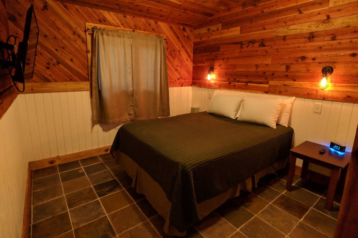 A wooden cabin-style room with a bed, a nightstand, a wall-mounted light, and a window with curtains.