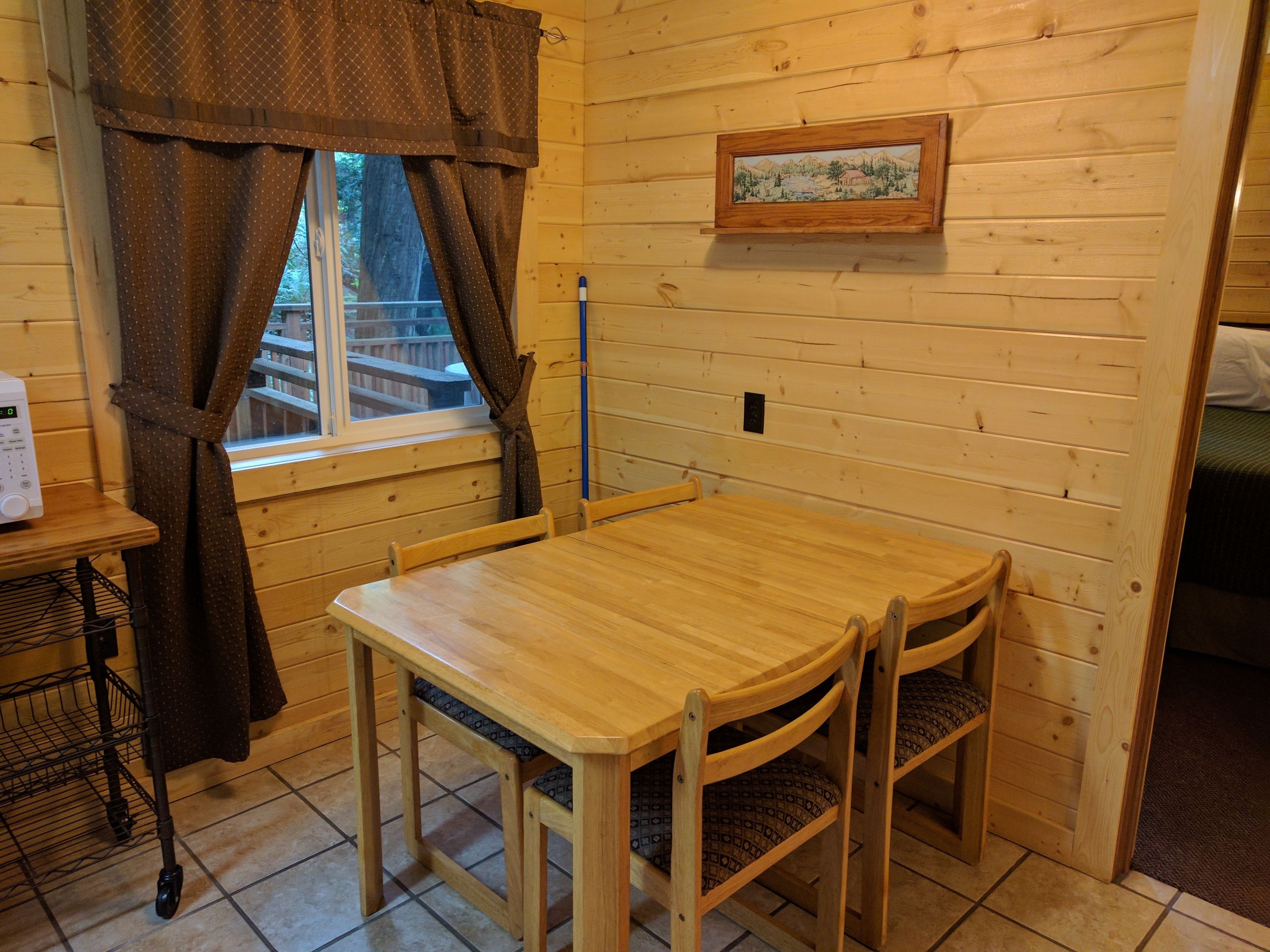 A wooden interior with a dining table and chairs, a window with curtains, and a framed picture on the wall.