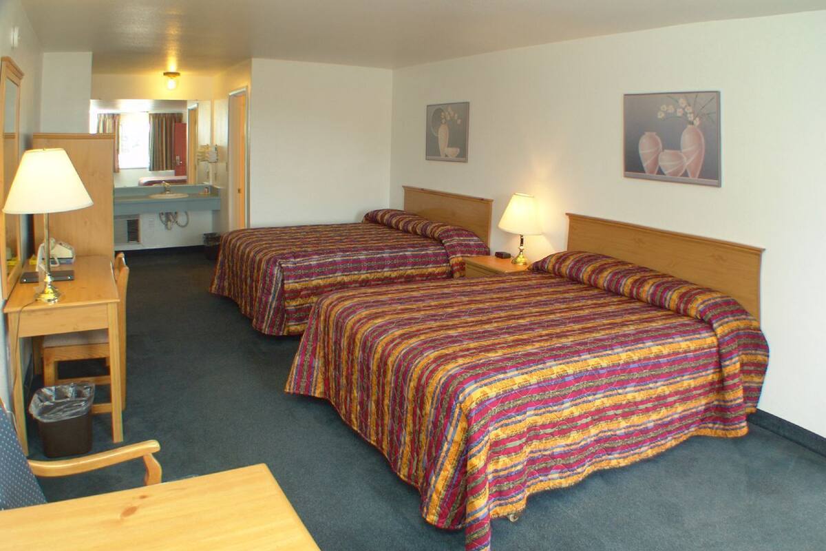 A hotel room with two beds, a desk, a chair, a lamp, and a trash bin.