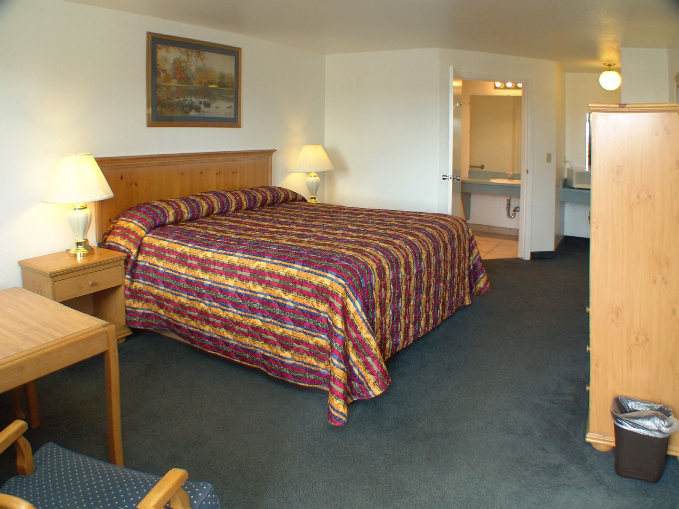 A hotel room with a bed, a desk, a chair, a lamp, a painting on the wall, and a bathroom visible through an open door.