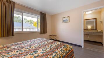 Une chambre d’hôtel avec un grand lit, une fenêtre avec des rideaux, une salle de bain avec un lavabo et un miroir, et un tableau accroché au mur.