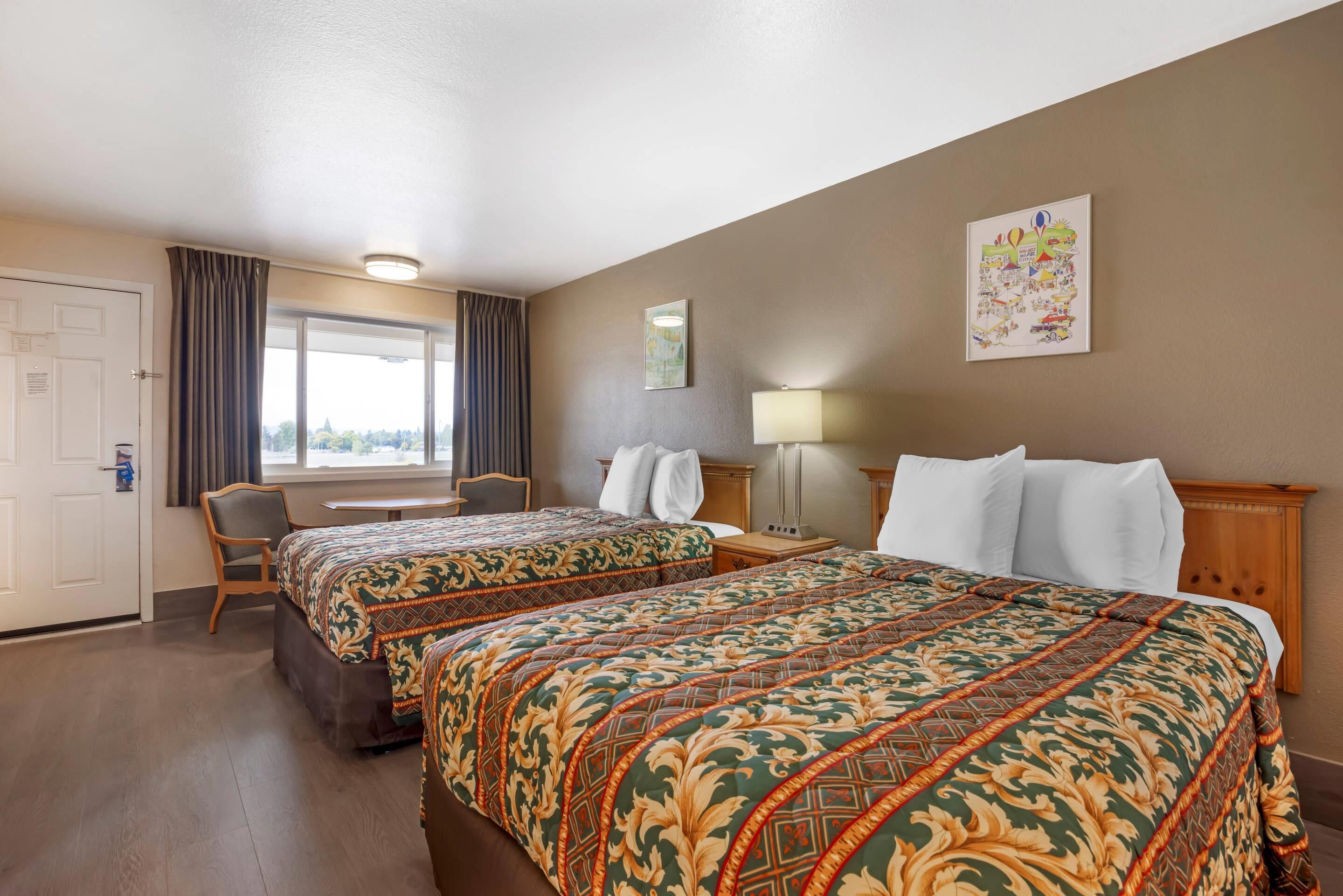 A hotel room with two beds, a desk, a chair, a window with curtains, a lamp, and a wall decoration.