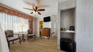 Habitación de hotel con piso de madera, ventilador de techo, televisor, cómoda, cafetera y basurero.