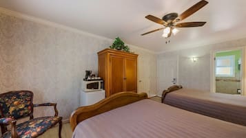 A bedroom with a bed, a wooden wardrobe, a microwave, a floral armchair, and a ceiling fan.
