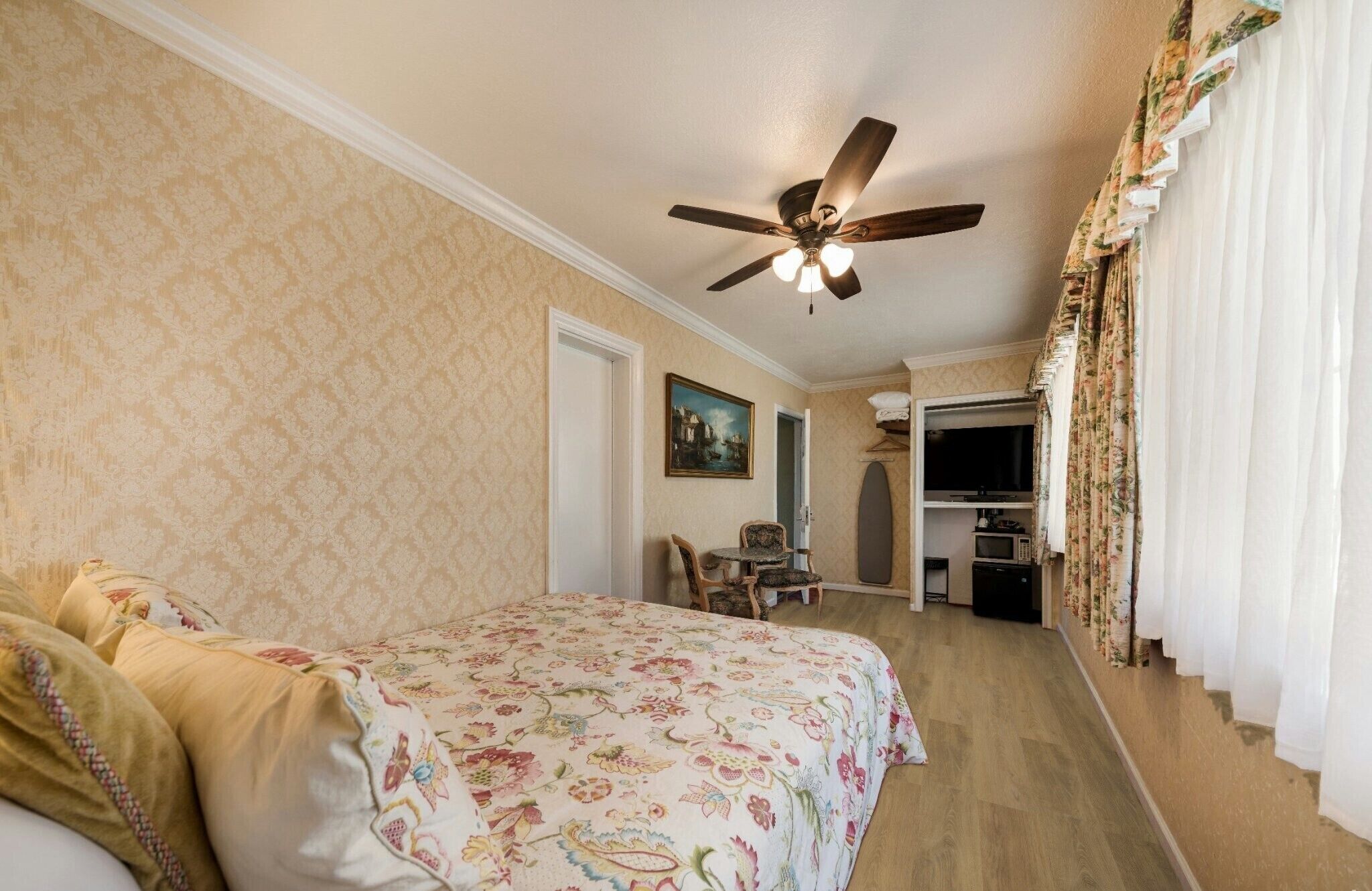 Une chambre d’hôtel avec un lit, un ventilateur de plafond, un tableau, une petite table et un coin cuisine.