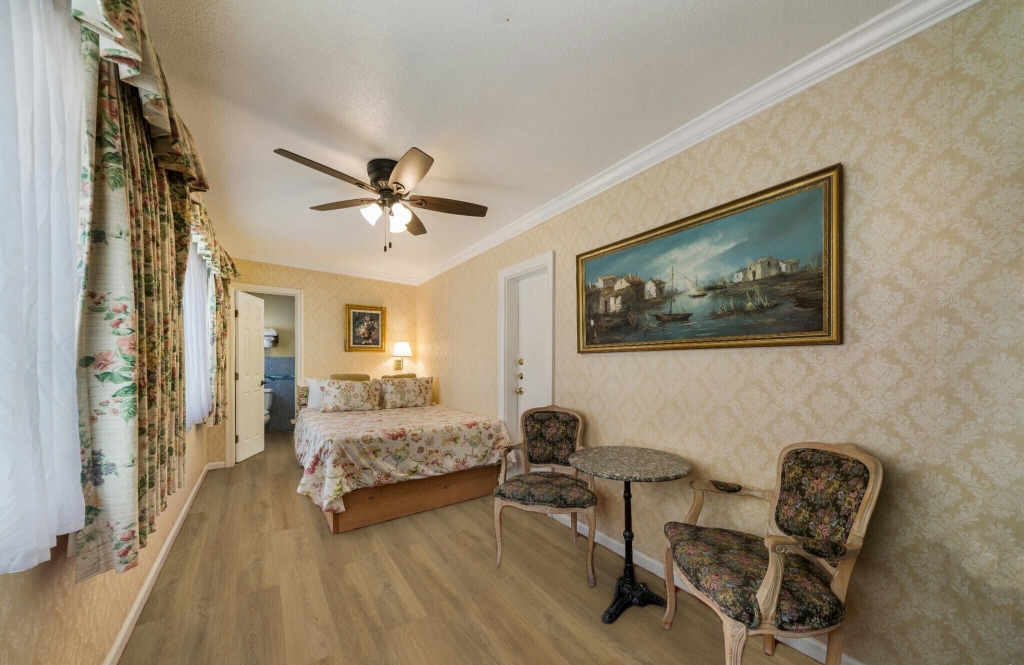 A hotel room with a bed, two chairs, a small table, a ceiling fan, a painting on the wall, and a door leading to a bathroom.