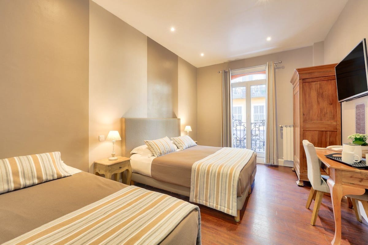 A hotel room with two beds, a wooden desk, a chair, a TV, and a balcony with a view.