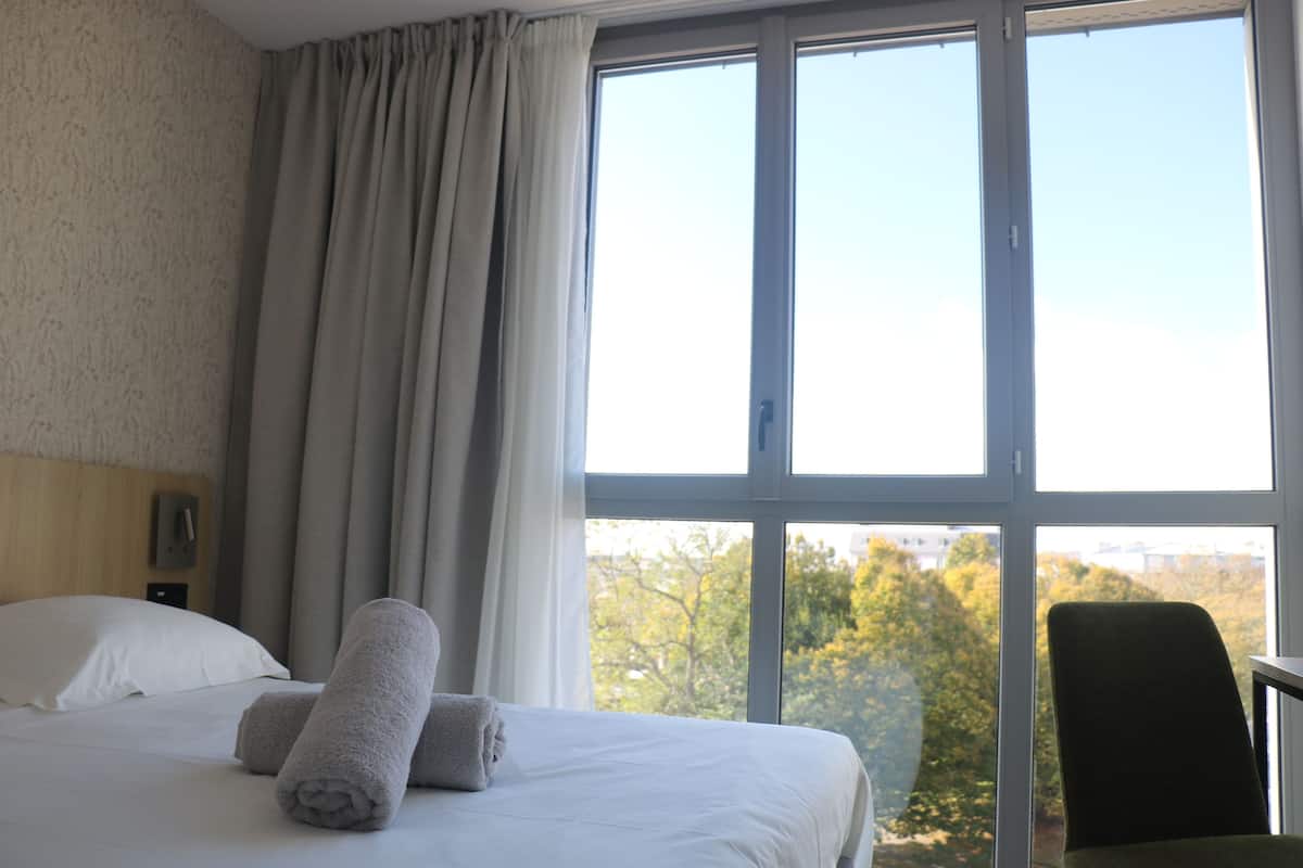 Una habitación de hotel con un ventanal grande, una cama con ropa blanca y una silla.
