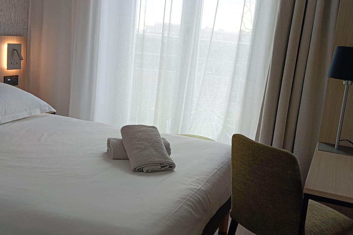Una habitación de hotel con una cama bien tendida, una silla, un escritorio y una ventana con cortinas translúcidas.