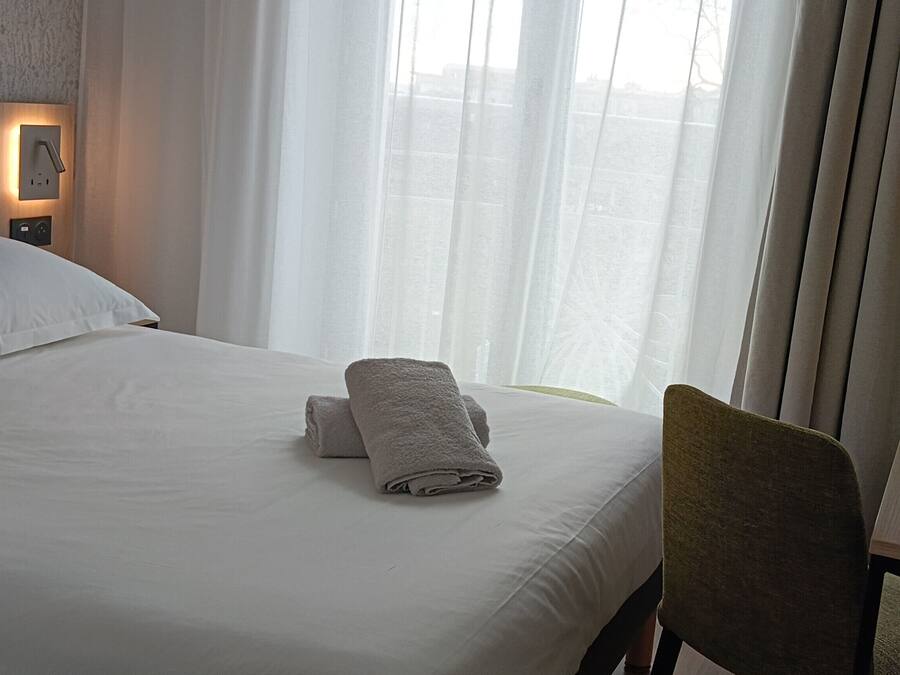 A hotel room with a neatly made bed, a chair, a desk, and a window with sheer curtains.