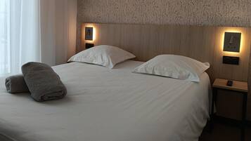 Una cama bien hecha con sábanas blancas y dos almohadas, una toalla gris y un cabecero de madera con luces fijadas a la pared.