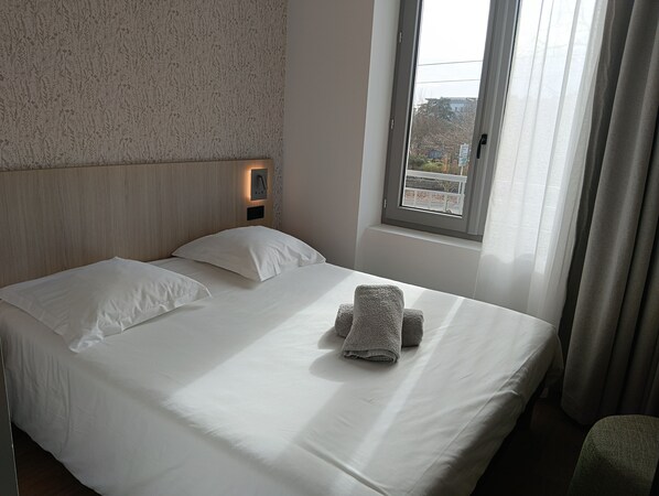 Una cama impecablemente hecha con sábanas blancas y dos toallas enrolladas, una lámpara de pared y una ventana con cortinas translúcidas.