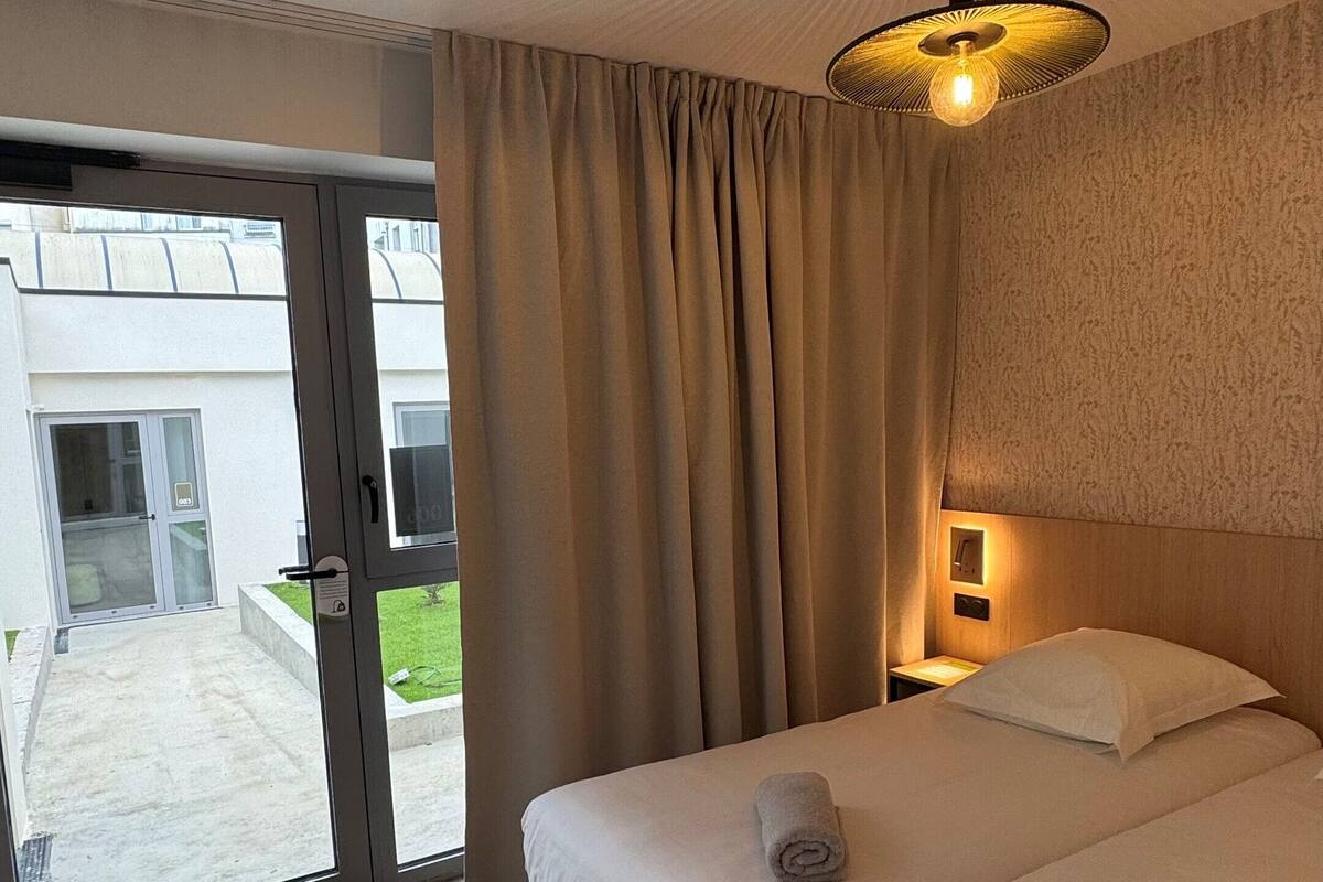 Habitación de hotel con dos camas, una puerta corrediza al exterior, un taburete pequeño verde y una lámpara en el techo.