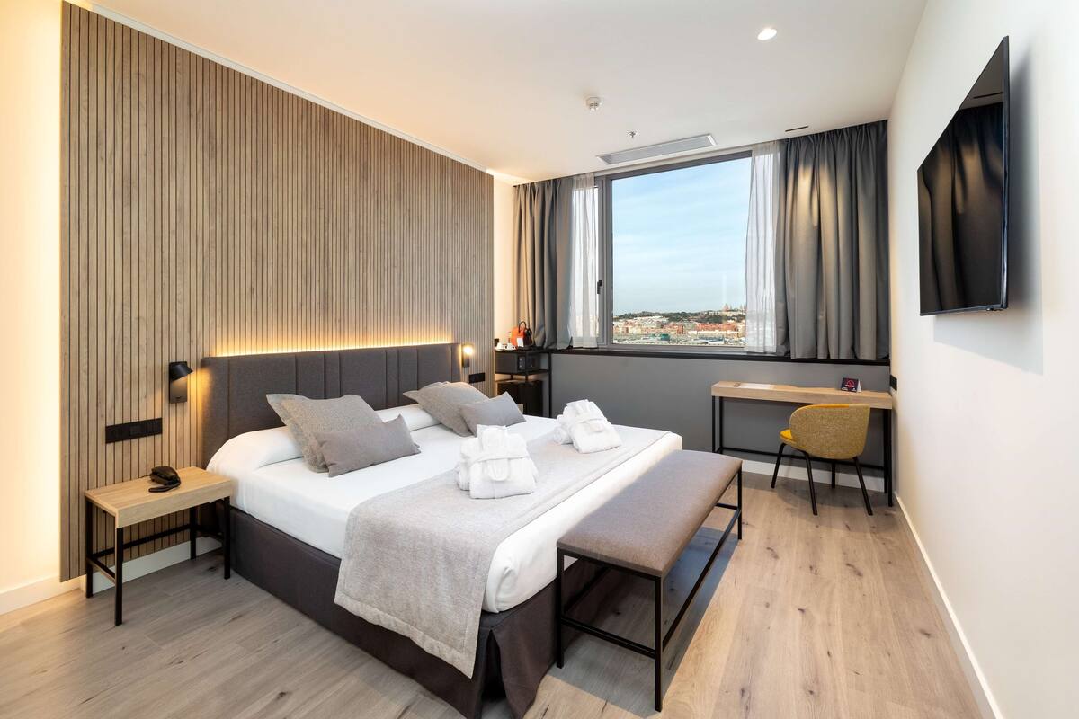 A modern hotel room with a large bed, bedside tables, a desk, a chair, and a TV mounted on the wall.