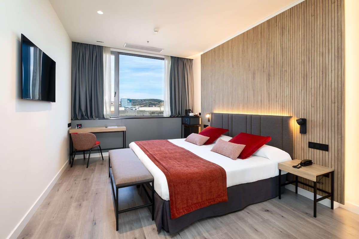 A modern hotel room with a bed, a desk, a chair, and a view of the city.