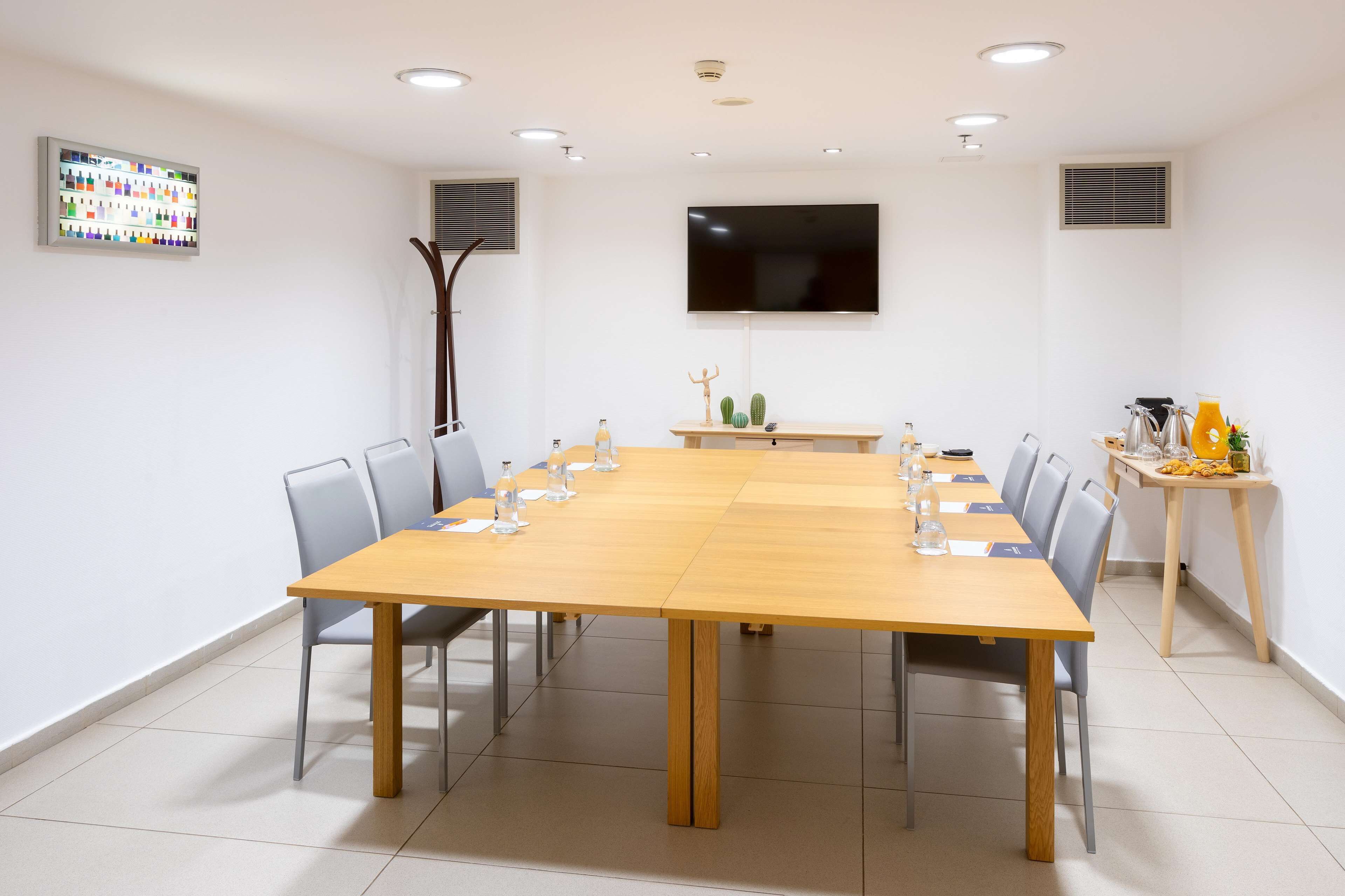 Un salón de conferencias con una larga mesa, sillas, un televisor montado en la pared y una pequeña mesa con una planta y una botella.