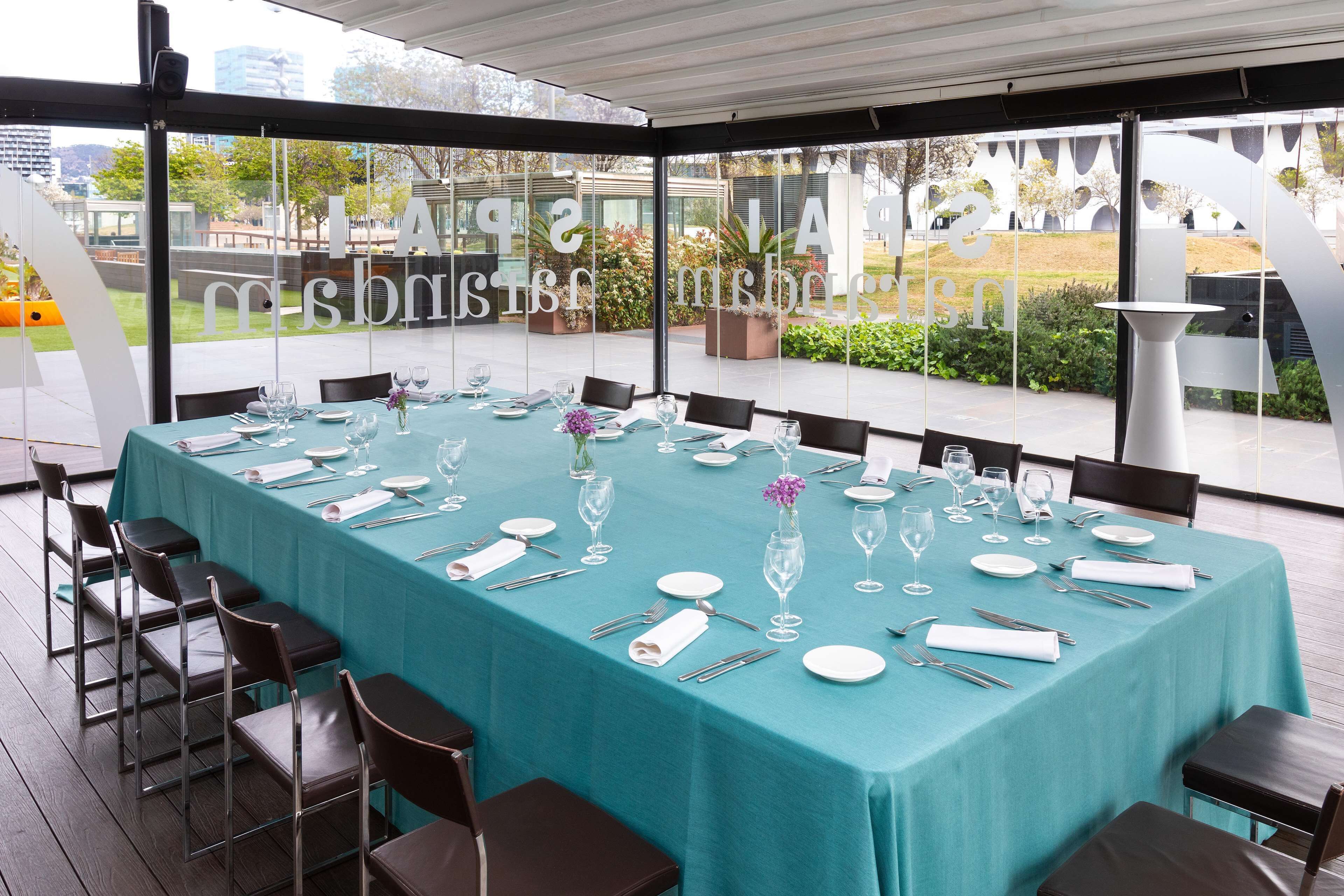 A large, rectangular table set for a formal event with chairs and glassware, under a covered area with large windows.