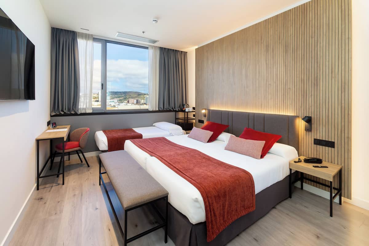A modern hotel room with a large bed, a desk, a chair, a bench, and a TV.