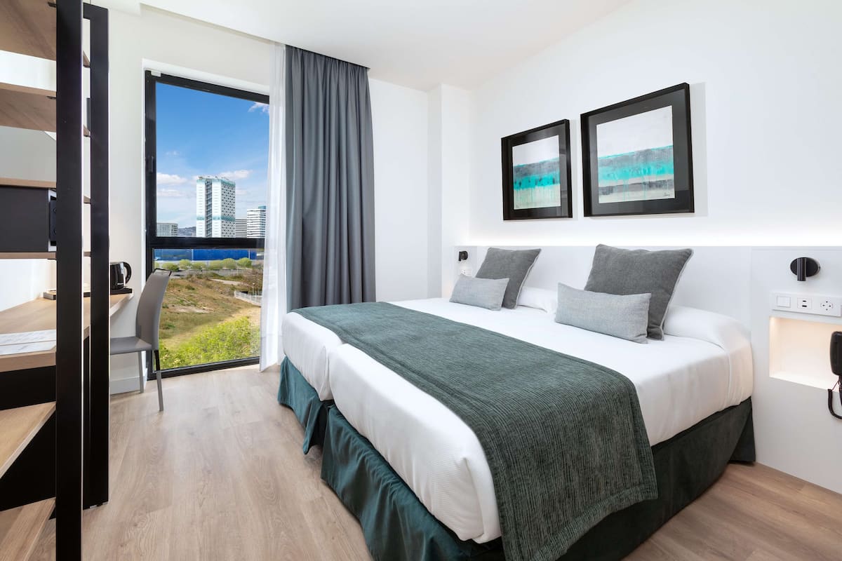 A modern hotel room with a large bed, a desk, and a view of a cityscape through a large window.