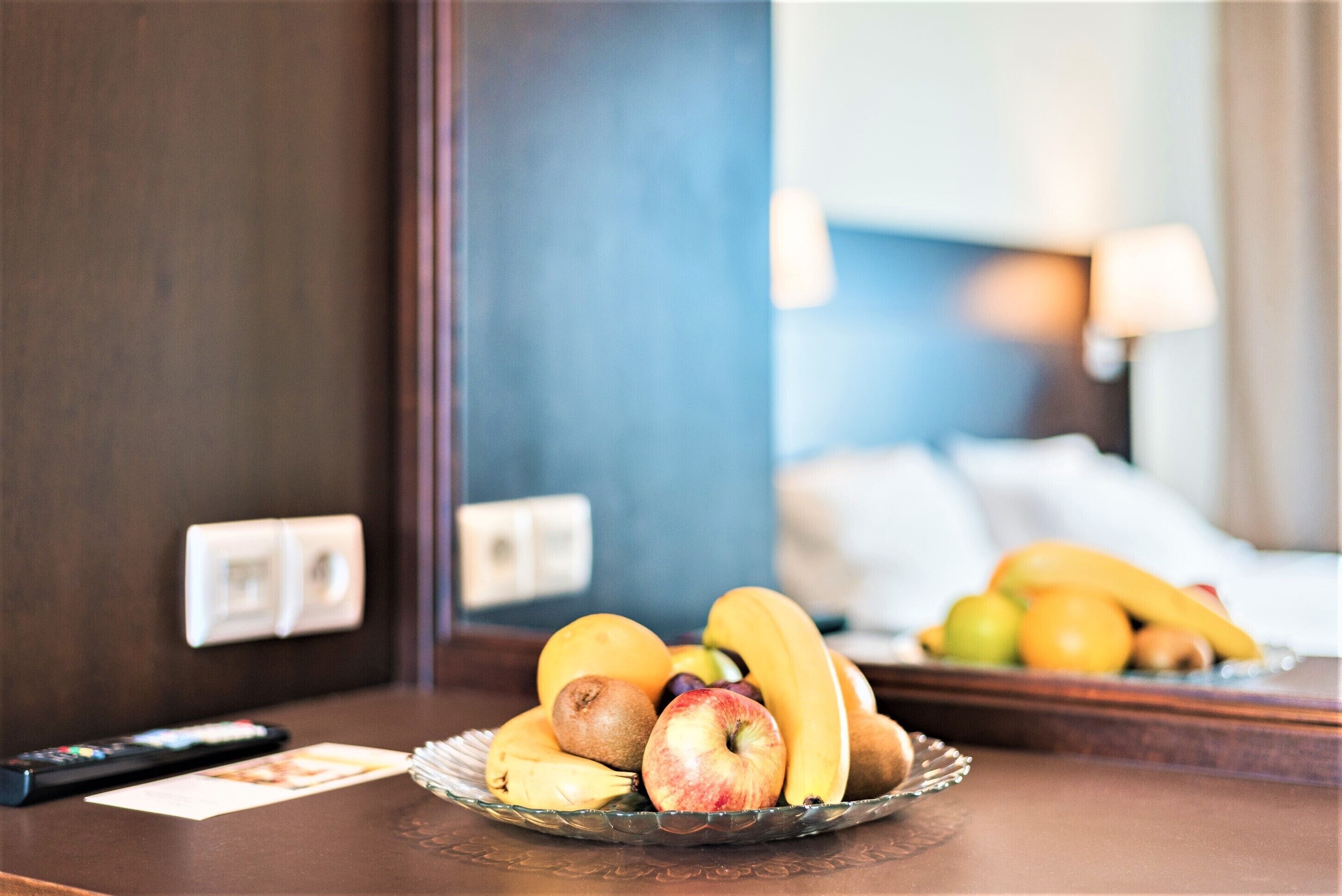 A hotel room with a fruit bowl on the table, a remote control, and a bed with a lamp in the background.